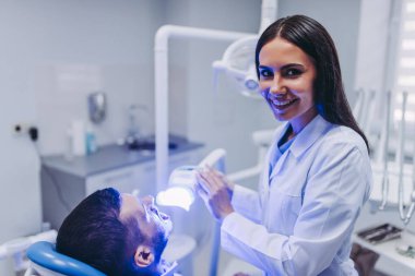 gülümseyen doktor hasta modern diş kliniği dişlerde tedavisi için uv lambası kullanırken kameraya bakıyor