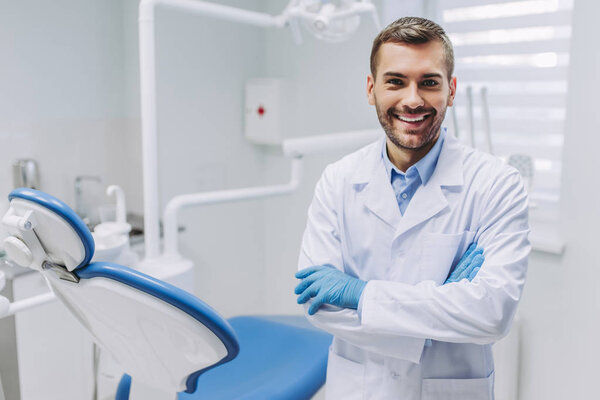 smiling doctor in medical gloves with crossed hands looking at the camera in dental clinic