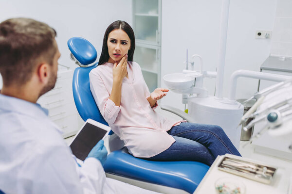 Doctor holding digital tablet talking with patient having toothache sitting in dental chair