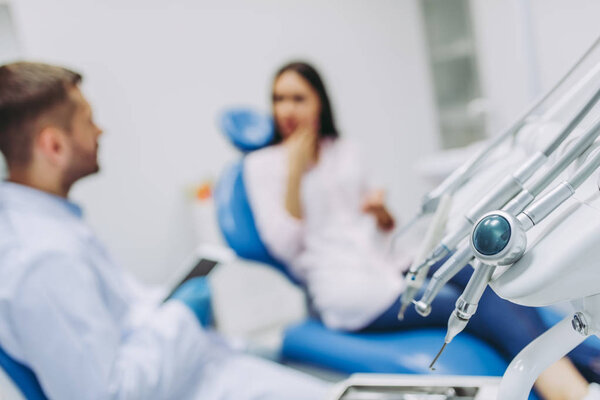 close up view of dental tools with doctor talking with patient having toothache sitting in dental chair on the background