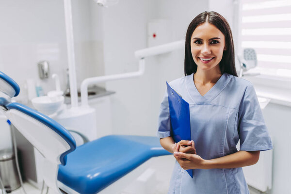 portrait of smiling female dentist with clipboard at working place in modern medical clinic