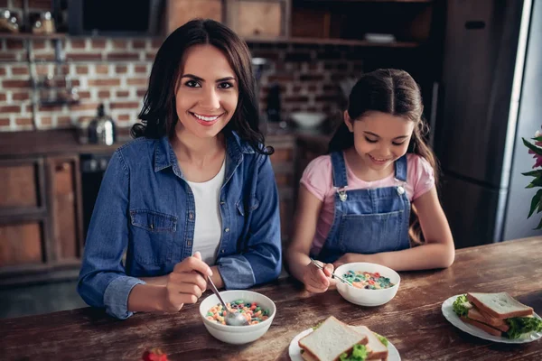 portrait of mother and daughter sitting at the table and eating colorful corn flakes for breakfast in the kitchen and looking at the camera