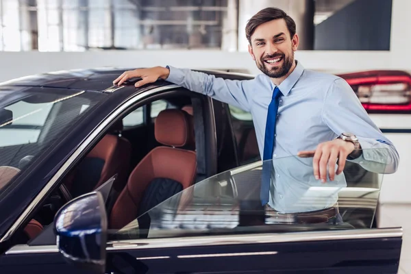 salesman standing at the car with opened door in dealership salon and looking at the camera