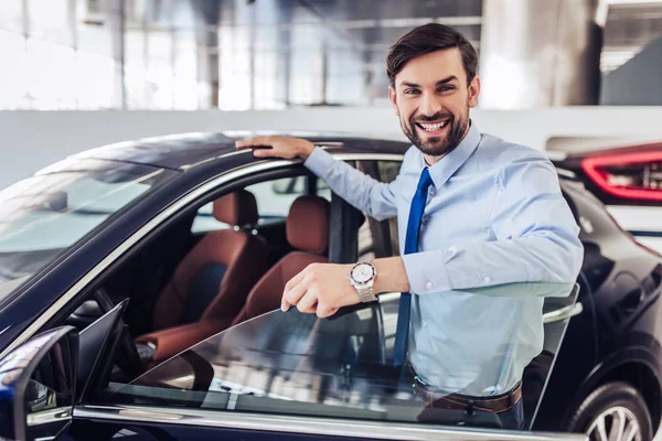 portrait of smiling salesman standing at the car with opened door in dealership salon and looking at the camera