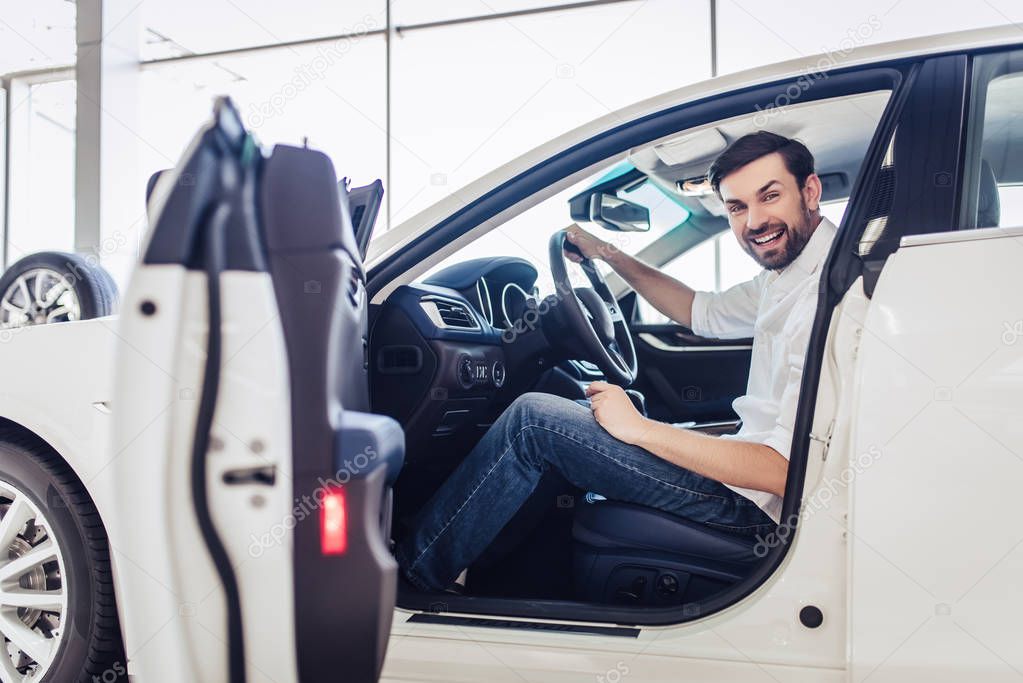 portrait of smiling bearded businessman sitting in the car with opened drivers door in dealership salon