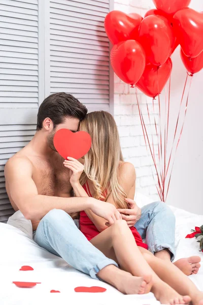 Jovem Casal Sentado Cama Fechando Rostos Com Coração Papel Vermelho — Fotografia de Stock