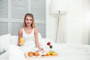 genç gülümseyen kadın holding cam el yatakta kahvaltı ile portakal suyu ve kameraya bakıyor