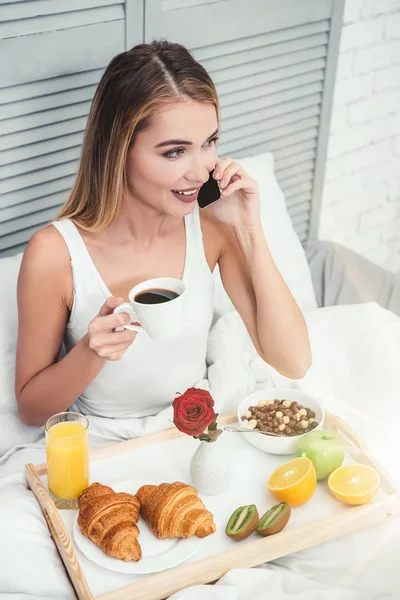 blonde woman talking on smartphone in the bed while drinking coffee