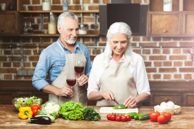 smiling man holding glasses with red wine while woman preparing salad at the kitchen
