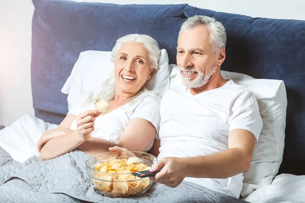 senior man and woman watching tv and eating chips in the bed and looking at the camera