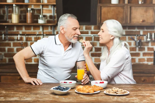 attractive senior woman feeding man with cookie at the kitchen