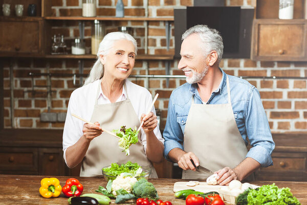 senior woman tossing salad and looking at the camera with man slicing vegetables
