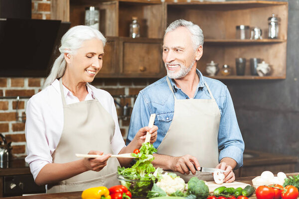 smiling senior man looking at woman preparing salad at the kitchen