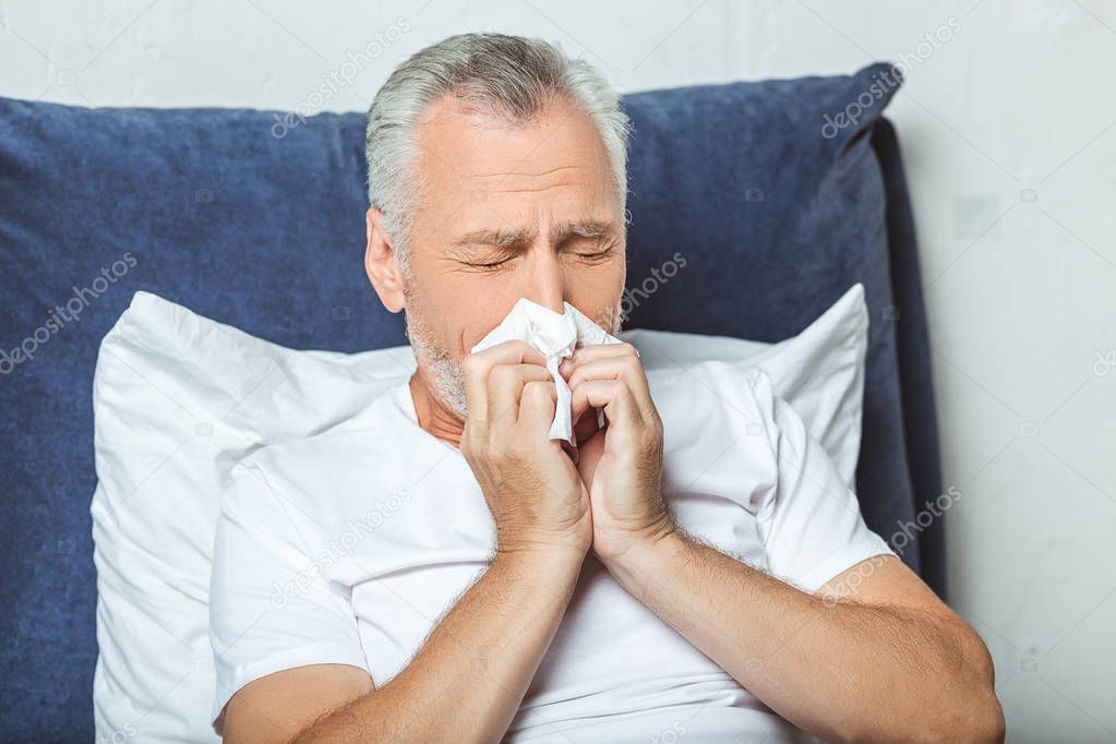 senior man sneezing in napkin sitting on the bed at home