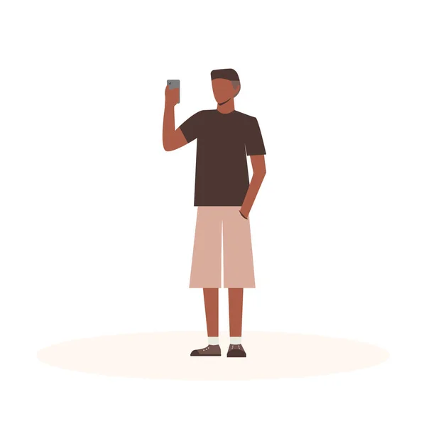 El hombre negro sostiene un teléfono y se toma selfie. Personaje plano de dibujos animados sobre fondo blanco aislado. Ilustración vectorial — Vector de stock