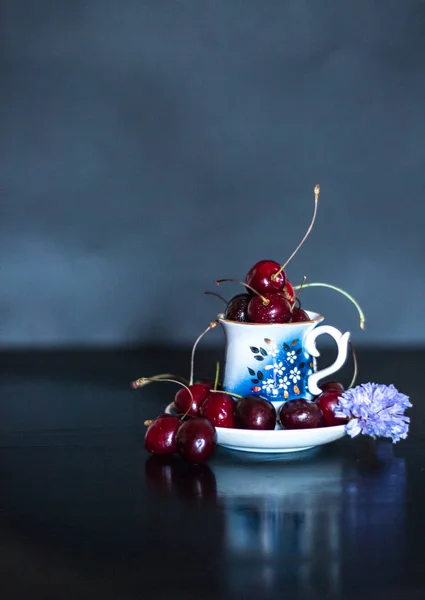 Donkere stillife met porseleinen kopje verse kersen met paarse bloem — Stockfoto