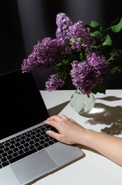 Cam vazoda parlak leylak dalıyla dolu bir hayat. Dizüstü bilgisayarı açık ve elleri gri perde arkasında masanın üzerinde.