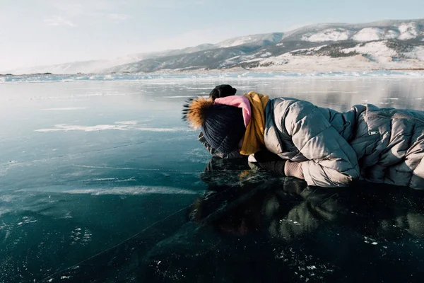 La ragazza giace sul ghiaccio e guarda attraverso il ghiaccio, esamina il fondo del lago Baikal. sotto il ghiaccio può essere visto passare da pesci e alghe sul fondo — Foto Stock