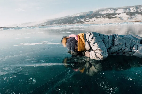 La ragazza giace sul ghiaccio e guarda attraverso il ghiaccio, esamina il fondo del lago Baikal. sotto il ghiaccio può essere visto passare da pesci e alghe sul fondo — Foto Stock