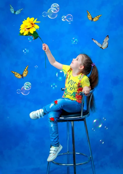 Estudio de rodaje. Una chica con un girasol en la mano sobre un fondo azul atrapa mariposas y pompas de jabón . —  Fotos de Stock