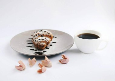 Kruvasan fincan kahve ve pembe çiçekler beyaz arka planda gri plaka üzerinde çikolata ve pudra şekeri ile