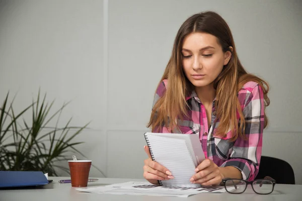 Junge Geschäftsfrau mit Brille in Freizeitkleidung, die am Tisch sitzt, schaut sich sorgfältig die Dokumente an — Stockfoto