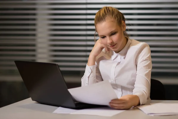 Junge Geschäftsfrau in lässiger Kleidung, am Tisch sitzend, Dokumente genau unter die Lupe nehmend, am Computer arbeitend — Stockfoto