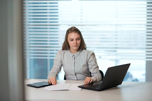 Junge Geschäftsfrau in lässiger Kleidung, am Tisch sitzend, Dokumente genau unter die Lupe nehmend, am Computer arbeitend — Stockfoto