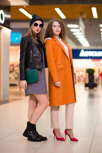 Beautiful girl with lush hair in a coat stands with the girl in jumpsuit and a black leather jacke indoors — Stock Photo, Image