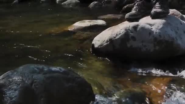 閉じる山の川を介して石の上を歩く人々の足のアップ 人々は互いに滑りやすい石の上を歩くのを助ける — ストック動画