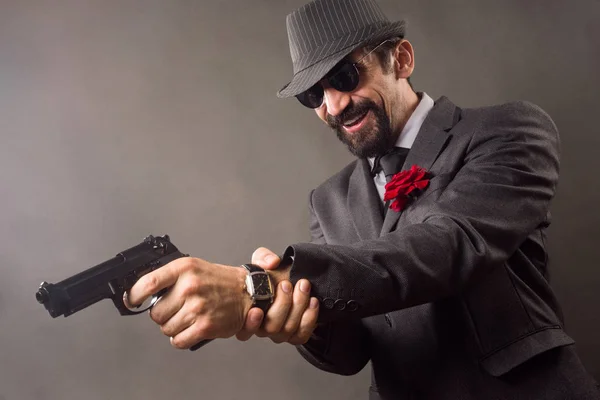 The smiling elegant gentleman in suit in classical retro style is aiming with pistol or handgun on gray background.