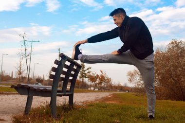 The sporty man is stretching on bench after jogging outdoors in park. clipart