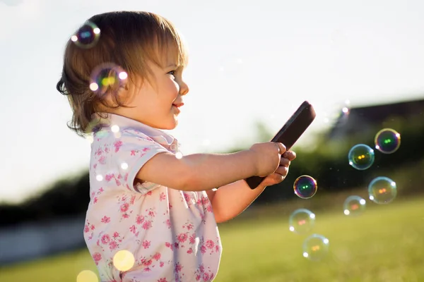 Het Schattige Babymeisje Met Behulp Van Telefoon Buitenshuis Met Zeepbellen — Stockfoto