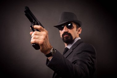 Şapka ve güneş gözlüğü ile takım elbiseli eski moda şık adam koyu gri arka plan üzerinde elinde tabanca tutuyor.