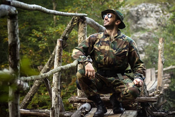 Smiling man in military uniform is squatting on the little bridge in nature, adventure in nature and orienteering concept.
