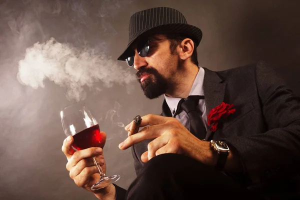 The elegant gentleman in suit with hat and sunglasses is smoking cigar and drinking red wine.