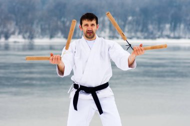 Orta yaşlı adam beyaz kimono ve siyah kuşak kış dövüş sanatları ile iki ahşap nunchaku pratik.
