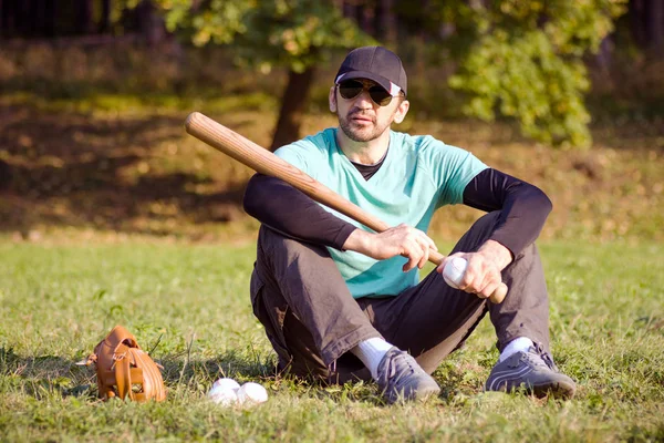 那个棒球运动员正坐在草地上 拿着他的运动器材休息 — 图库照片