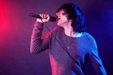 Genç adam vokalist mikrofon sahnede konser kırmızı ve mavi ışıklarla aydınlatılmış çığlık atıyor.