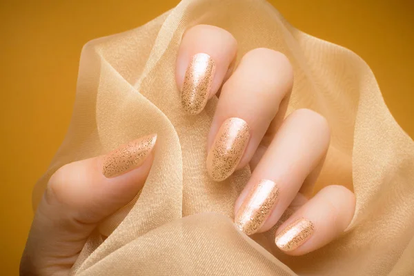 Female hand with glittered beige nails is holding beige textile on orange background.