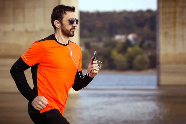 オレンジ色のスポーツ ウエアでフィット男はイヤホンと携帯電話 レクリエーションの概念と屋外ジョギングします ストック写真