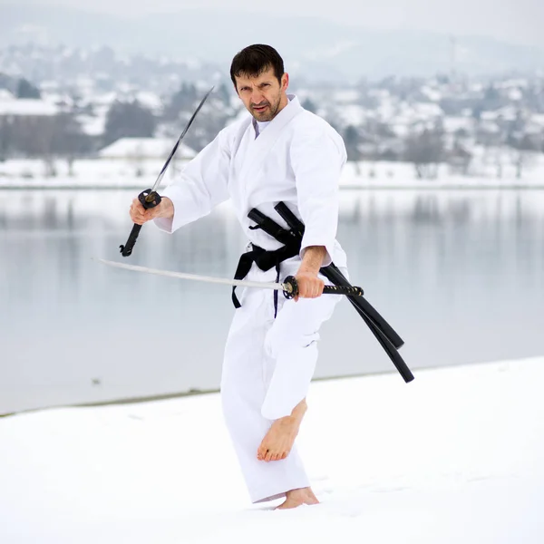 Man in white kimono is practicing martial arts with two samurai swords barefoot on snow at winter.