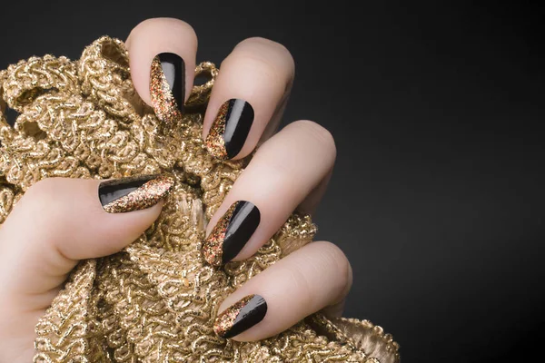 Female hand with black and golden glittered nails is holding golden decoration on black background, manicure and nail care concept.