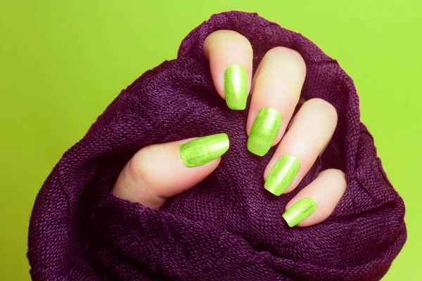 The female hand with neon green nails is wearing a purple fabric on green background, manicure and nail care concept.