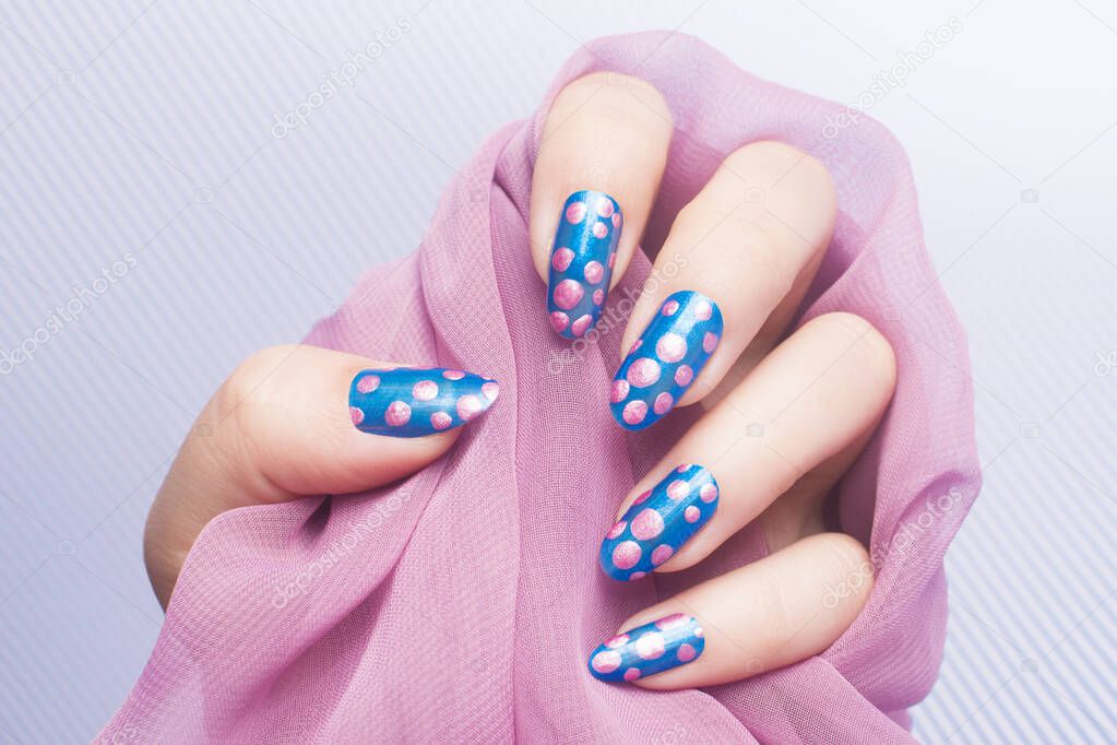 Female hand with pink spotted blue nails is holding pink fabric on striped background, manicure and nail art concept.