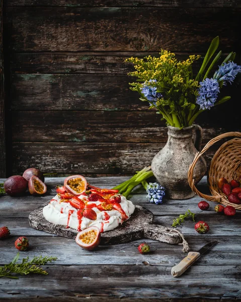 Yummy strawberry and passion fruit meringue cake on wooden board, vase with flowers on table — Stock Photo