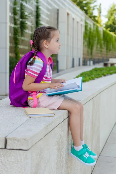 First day of school. Pupil of primary school with book in hand. . Girl with a backpack near the building  outdoor. Beginning of classes. The first day of autumn. concept back to school.