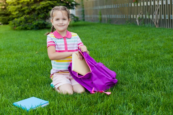 First day of school. child girl schoolgirl elementary school student sitting on the grass near the school and puts books in his backpack. Concept back to school. outdoor activities
