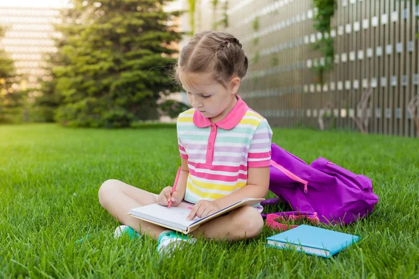 First day of school. child girl schoolgirl elementary school student sitting on the grass near the school and draws in a notebook. Concept back to school. outdoor activities