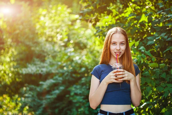 Genç güzel kız sulu yeşillik bir arka plan üzerinde taze sıkılmış meyve suyu içecekler.
